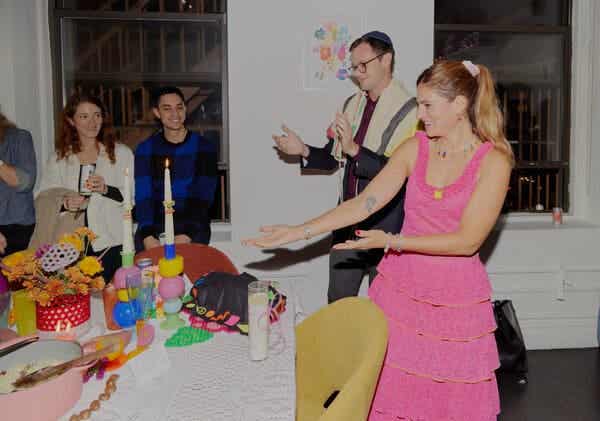 Susan Korn, wearing a tiered pink dress, gestures toward a Shabbat table while guests smile and Rabbi Matt Green applauds.