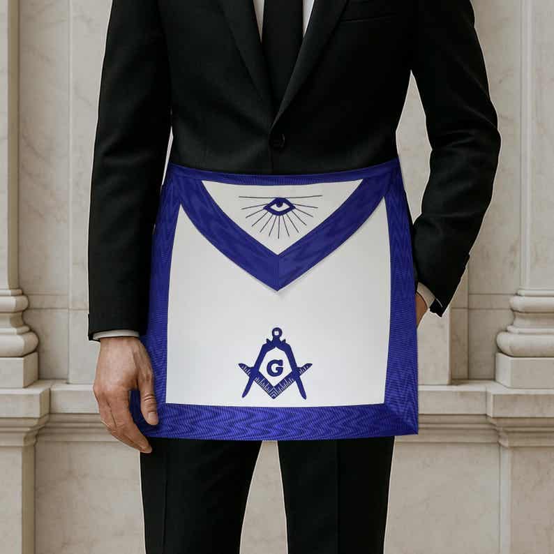 May include: A white apron with a blue border and accents. The apron features a design with an eye, sun rays, a compass, and a square. The apron is worn over a black suit.